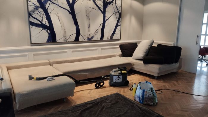 Un sofá modular color crema está siendo limpiado a fondo en una sala de estar, con un equipo de aspiración y una botella rociadora en el suelo.