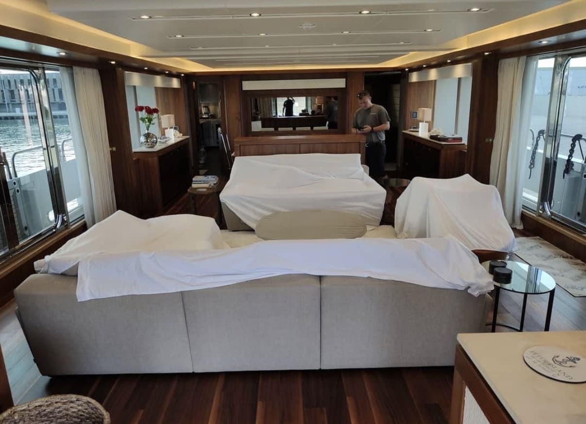 Interior de la zona de estar de un yate de lujo con muebles tapizados, suelo de madera y grandes ventanales con vistas al agua.