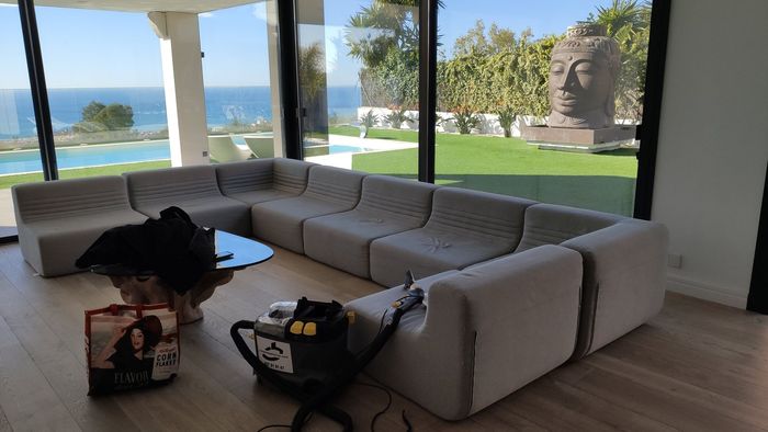 Un sofá modular de color beige está situado frente a una gran ventana con vistas a una piscina y al océano, con utensilios de limpieza en el suelo, cerca de allí.