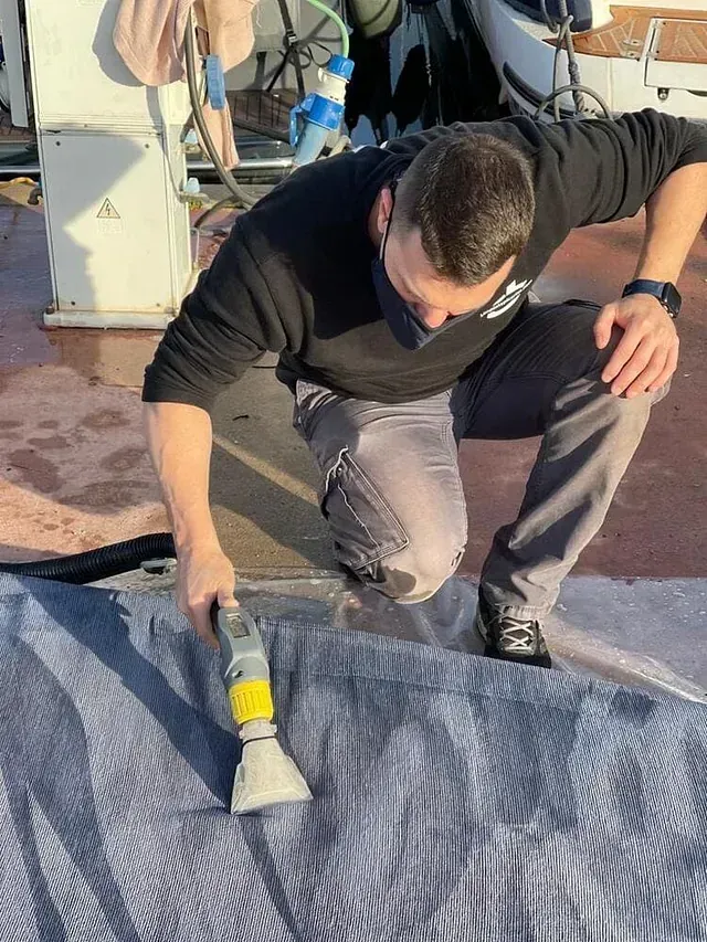 Una persona vestida con ropa oscura está arrodillada en un muelle, limpiando una lona gris para barcos con una herramienta de extracción.