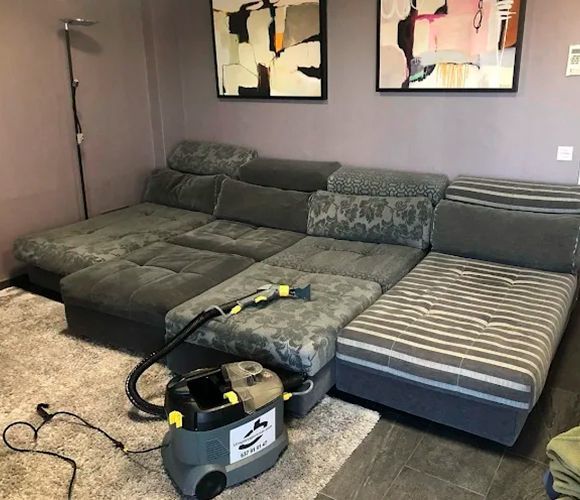 Limpiador de tapicería aspirando un gran sofá seccional gris en una habitación con obras de arte abstractas.