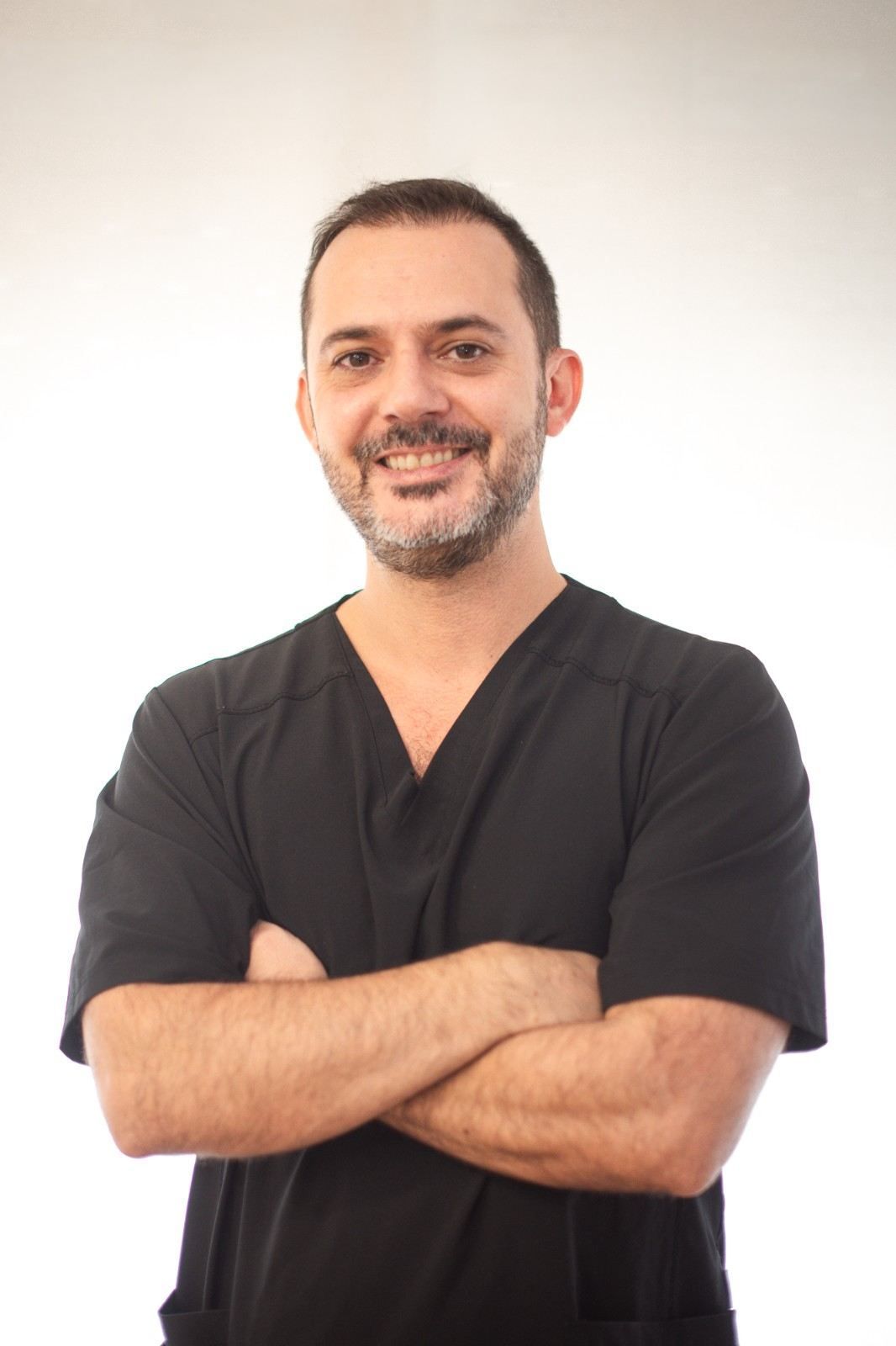 Miembro del personal sonriente con uniforme médico azul marino, gafas y un gran collar con colgante sobre un fondo blanco.