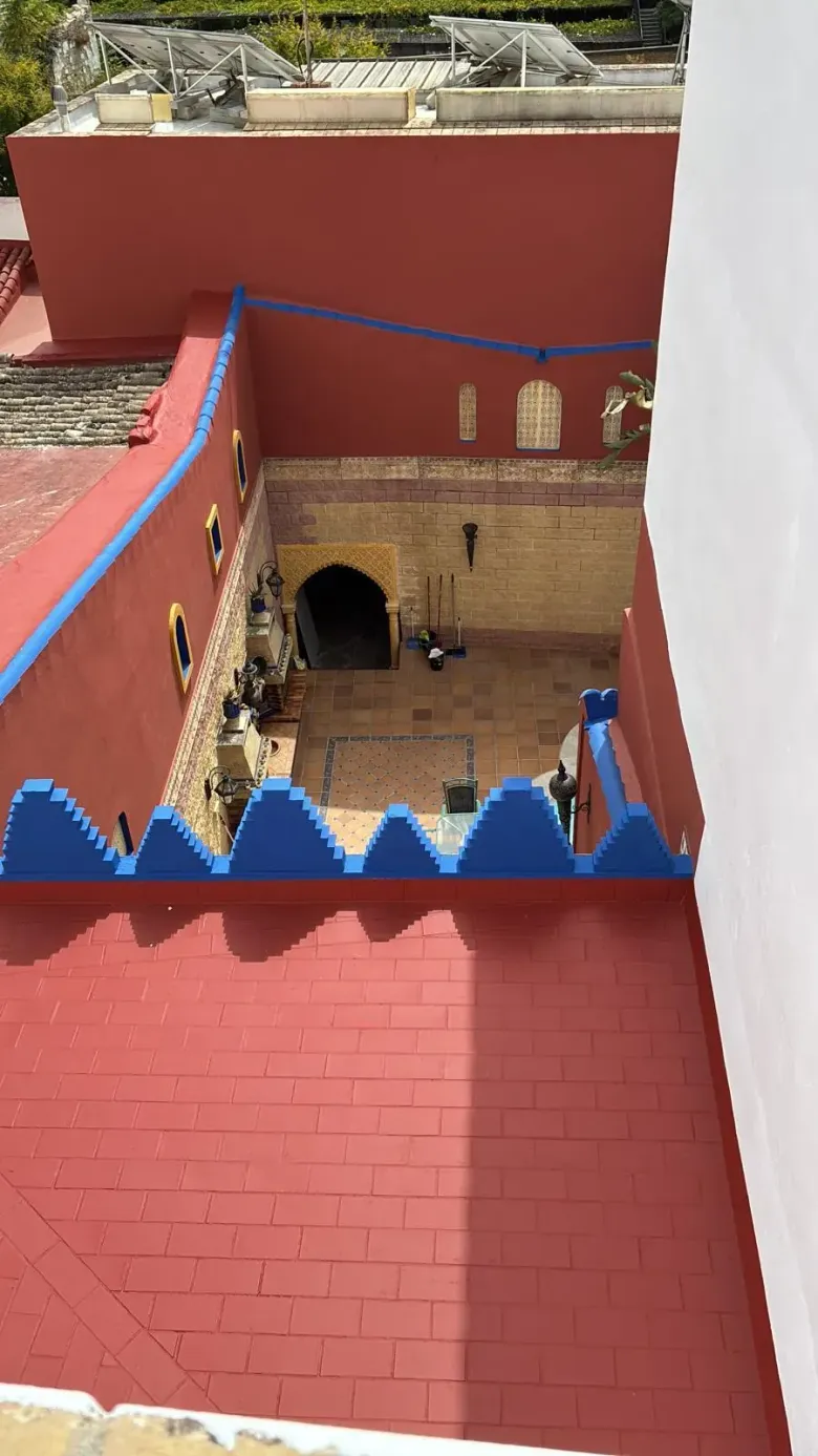 Exterior de edificio rojo y azul con patio. Ladrillo, arco y almenas azules. Día soleado.