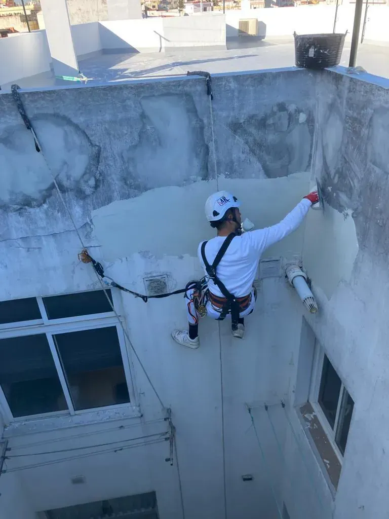 Hombre con arnés de seguridad aplicando pintura al exterior de un edificio. Paredes claras, día soleado.
