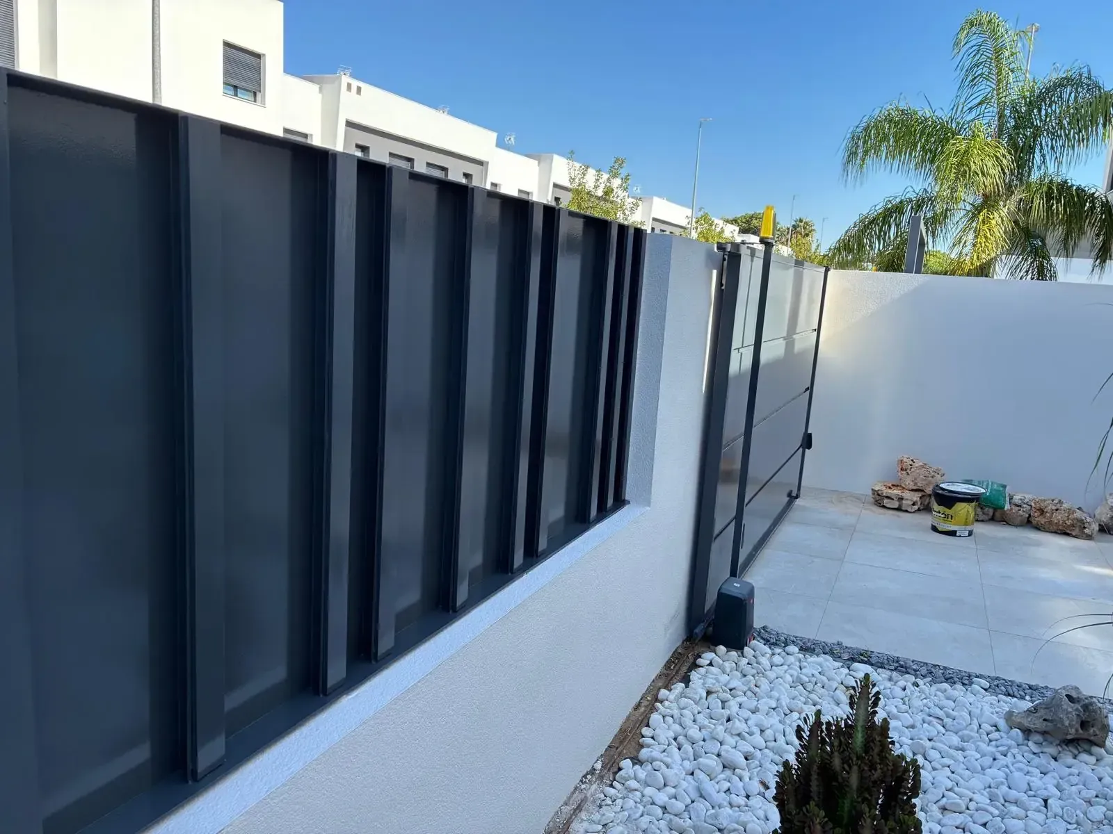 Valla y portón de metal gris sobre pared blanca, con pequeñas piedras y plantas al frente.