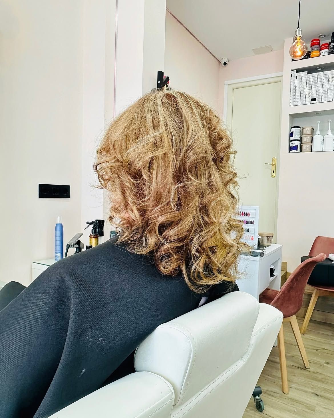 Una persona de cabello rubio y rizado está sentada en una silla blanca en un salón, de espaldas a la cámara.