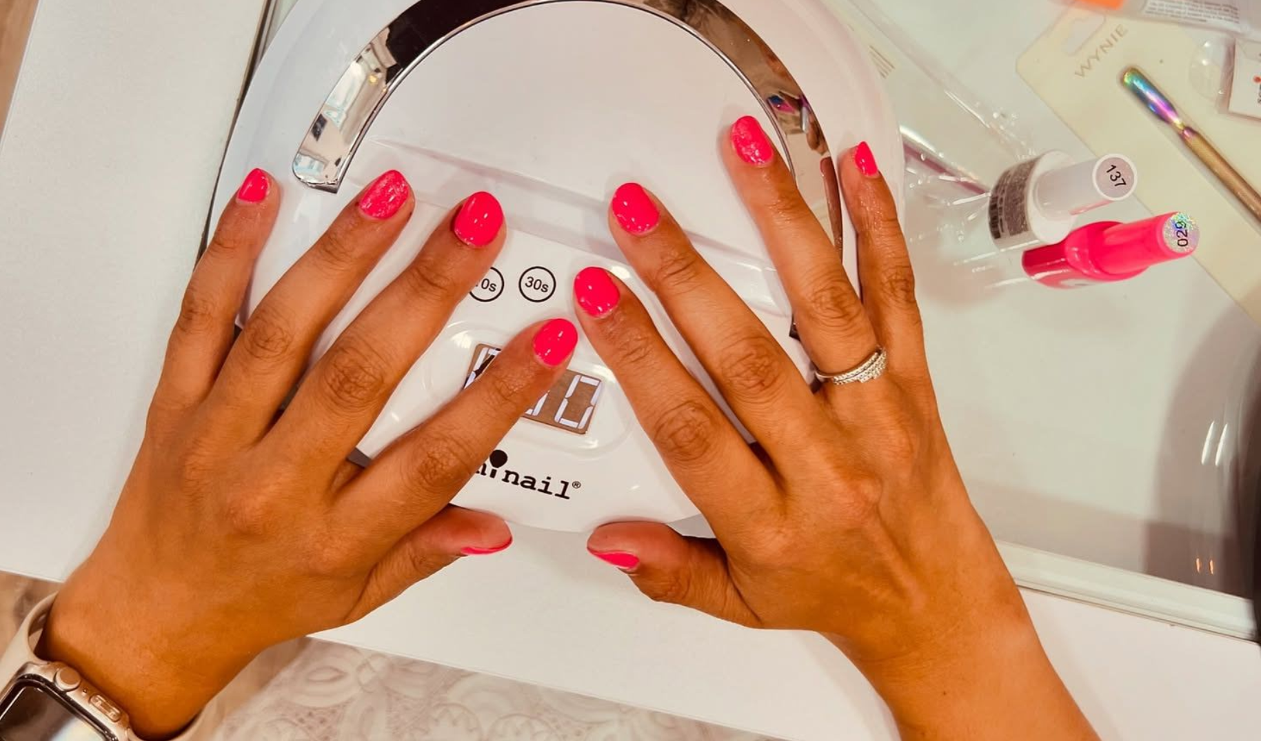 Manos con uñas de color rosa intenso sosteniendo un espejo redondo junto a unos frascos de esmalte de uñas.
