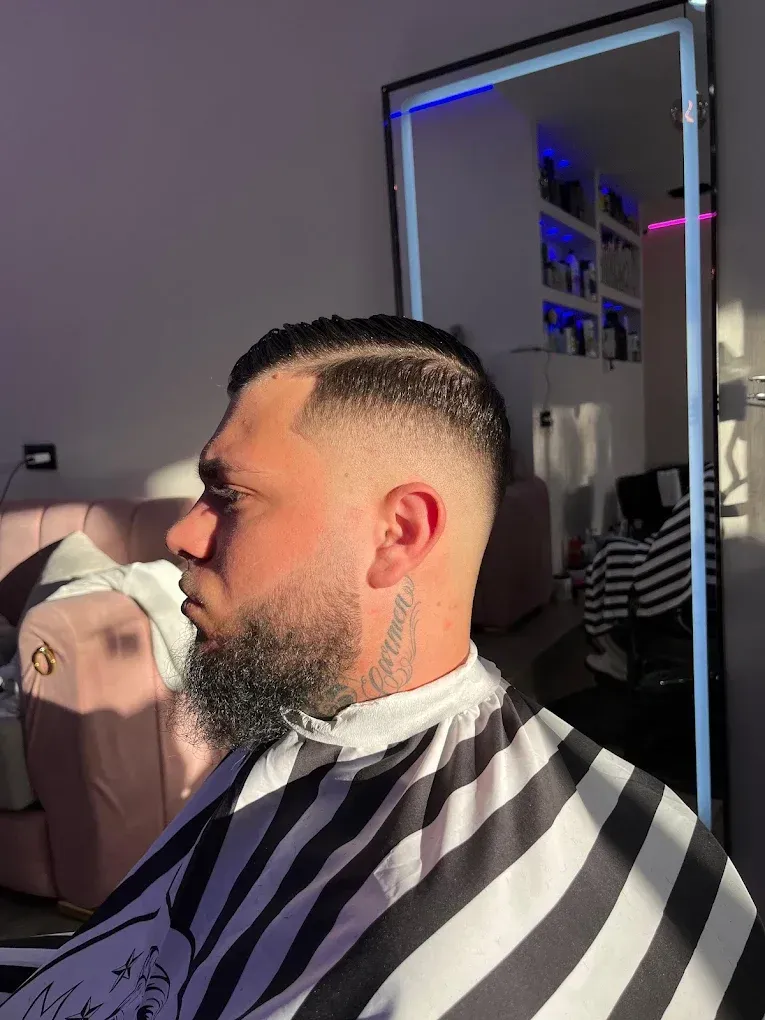 Hombre con una capa de peluquería a rayas y un degradado recién hecho, sentado en una silla de salón cerca de un espejo.
