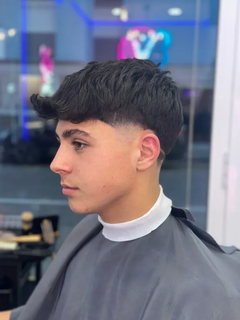 Persona joven con un corte de pelo oscuro y texturizado, sentado en una silla de barbero de perfil, con una capa negra