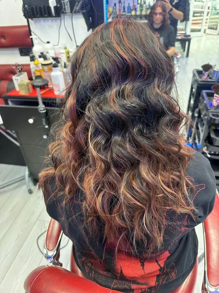 Cabello castaño largo y ondulado con mechas, sentada en una silla de peluquería, vista desde atrás