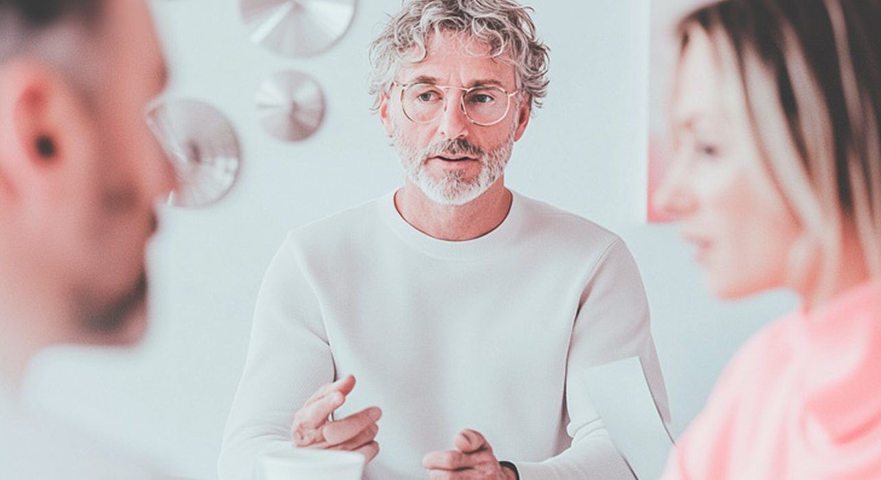 Valkoiseen neuleeseen pukeutunut ammattilainen puhuu ja elehtii istuen kahden muun henkilön vastapäätä valoisassa ja modernissa ympäristössä.