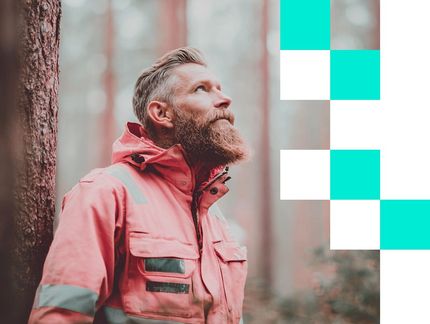 Parrakas henkilö, jolla on vaaleanpunainen takki, katsoo ylöspäin nojaten puuhun metsässä.
