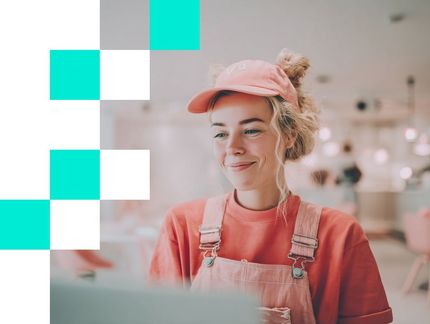 Henkilö, jolla on vaaleanpunainen lippalakki ja haalarit, hymyilee ja katsoo kannettavaa tietokonetta kirkkaassa, sumeassa sisätilassa.