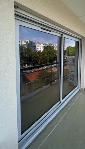 Fenêtre coulissante à vitrage réfléchissant sur un mur extérieur beige, donnant sur les arbres et les bâtiments.