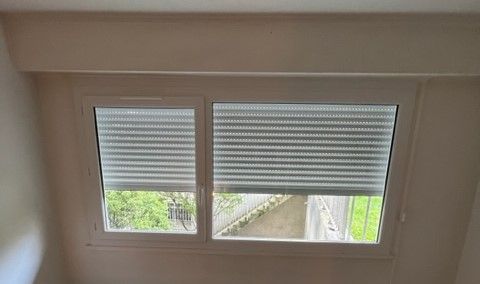 Fenêtre avec deux stores enrouleurs blancs, dont l'un est partiellement relevé, laissant apparaître la verdure à l'extérieur.
