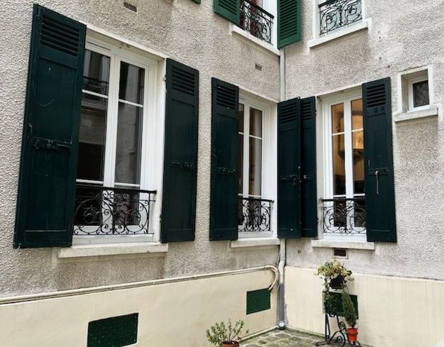 Une fenêtre blanche avec des volets bleu foncé et une petite rambarde en fer forgé noir, adossée à un mur de pierre claire.
