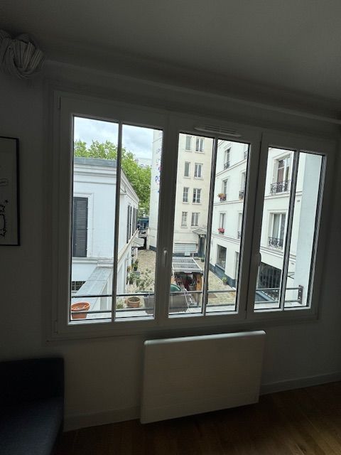 Vue à travers une fenêtre à cadre blanc sur des immeubles et une rue en contrebas, avec un radiateur en dessous.