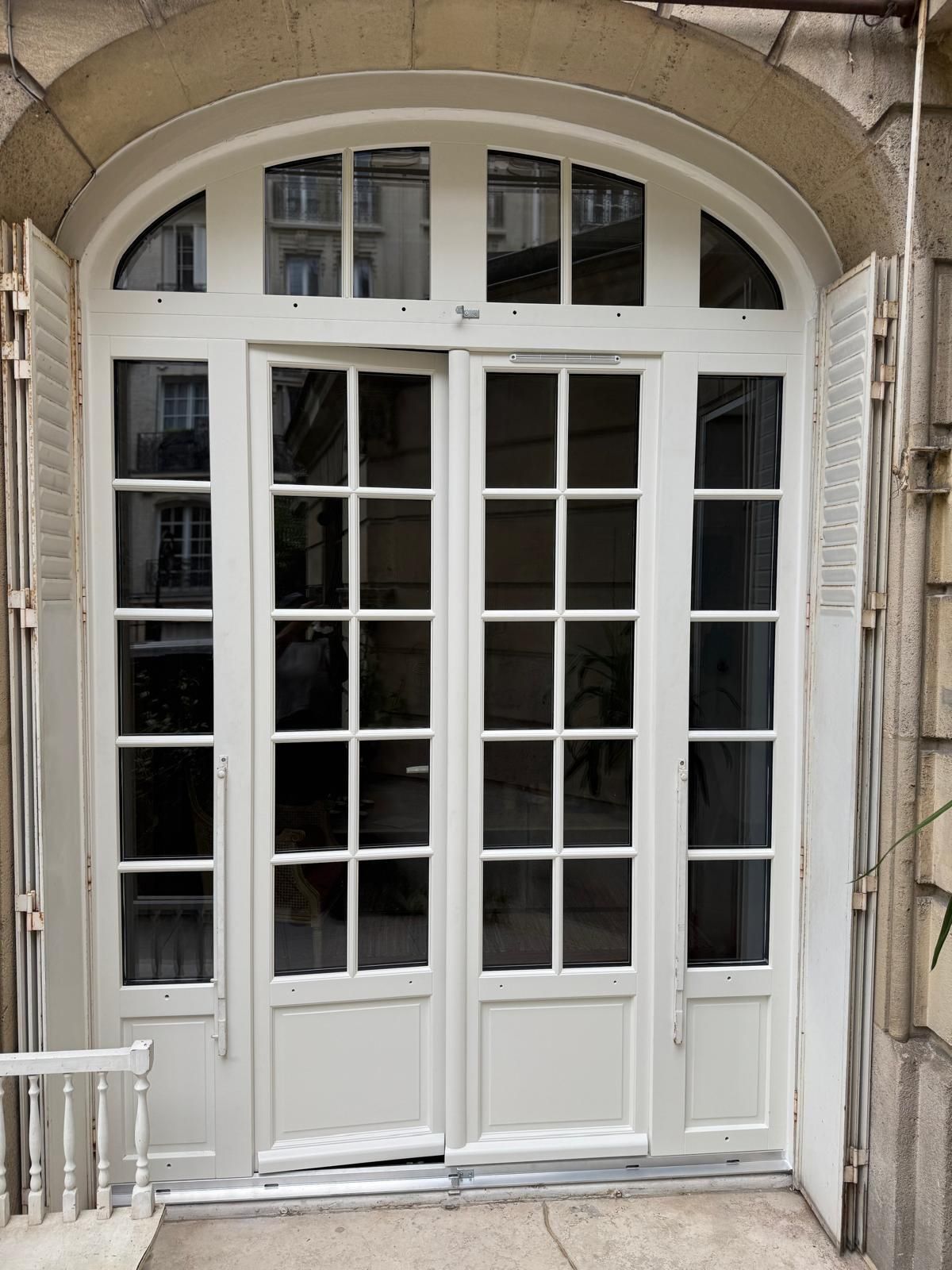 Double porte cintrée blanche à plusieurs panneaux de verre et fenêtres latérales, intégrée à une façade d'immeuble.