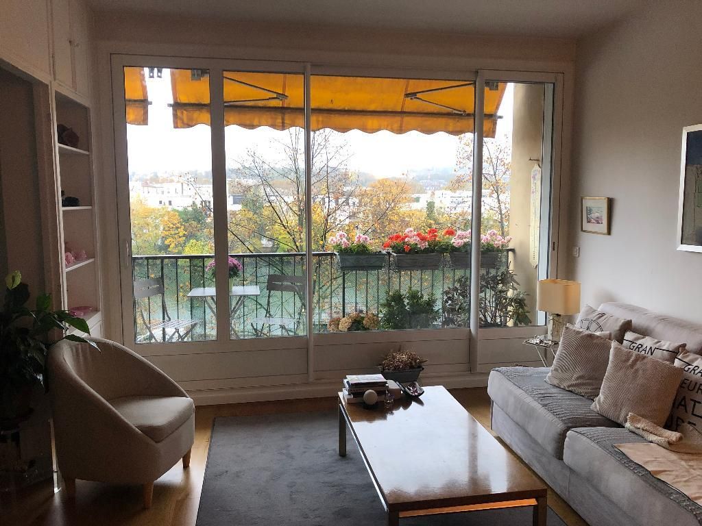 Un salon avec un canapé beige, un tapis gris et un fauteuil, doté d'une grande fenêtre donnant sur un balcon.