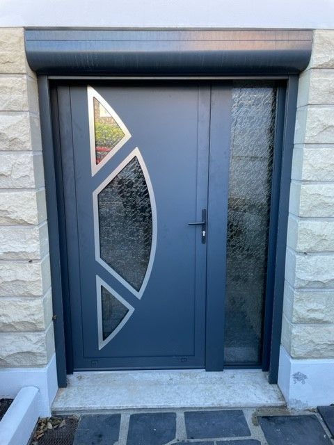 Porte d'entrée moderne grise avec panneaux en verre dépoli et motif géométrique ajouré sur façade en pierre