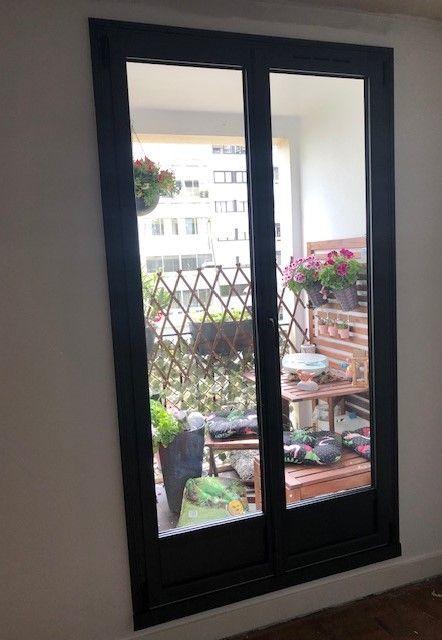 Porte-fenêtre à cadre noir donnant sur un petit balcon fleuri avec vue sur la ville