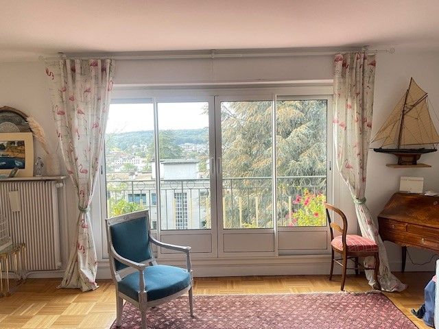 Salon cosy avec fauteuil bleu, grande fenêtre, rideaux à fleurs et petit décor de voilier.