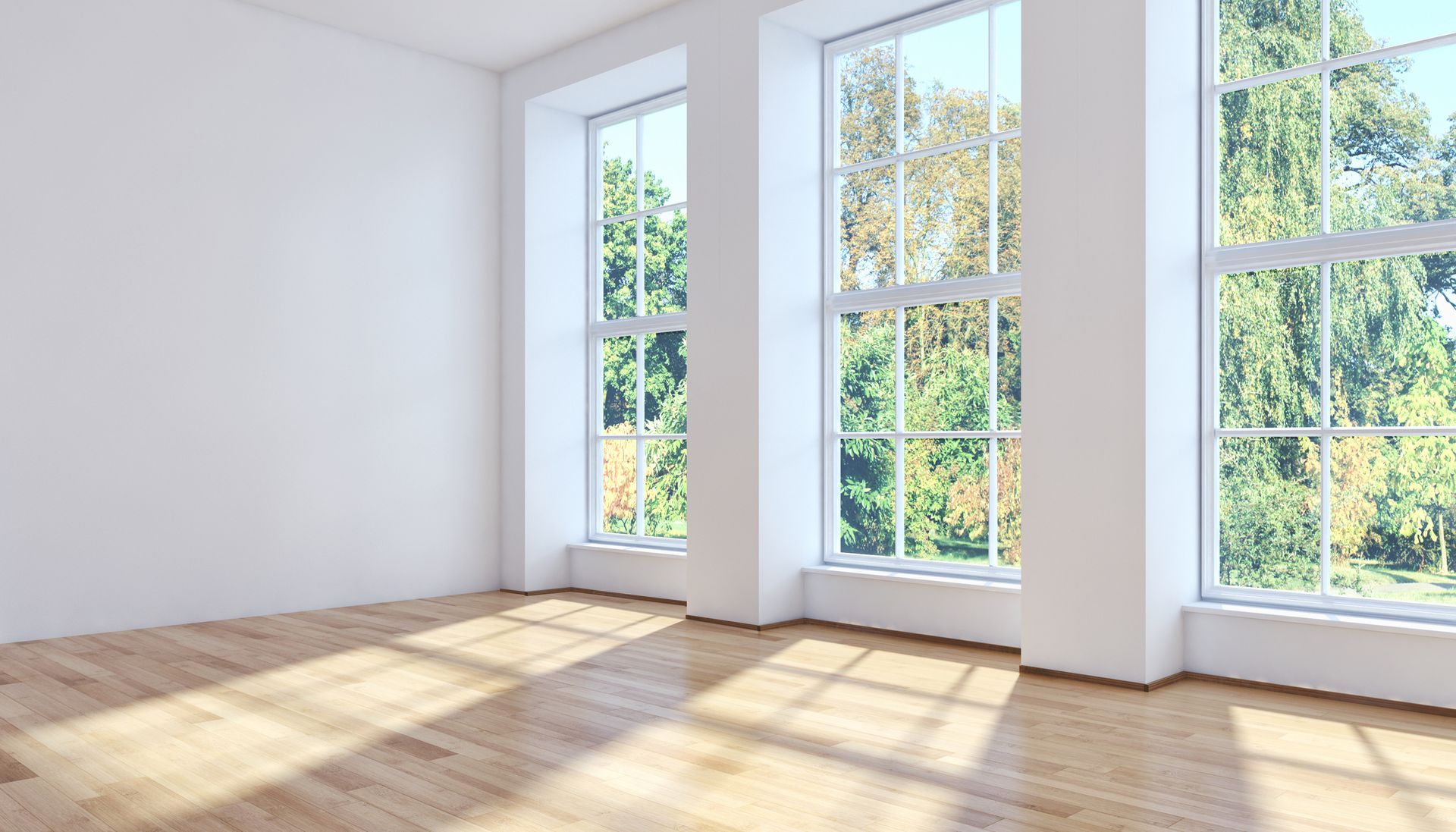 Une pièce vide avec un parquet clair, des murs blancs et trois hautes fenêtres donnant sur des arbres.
