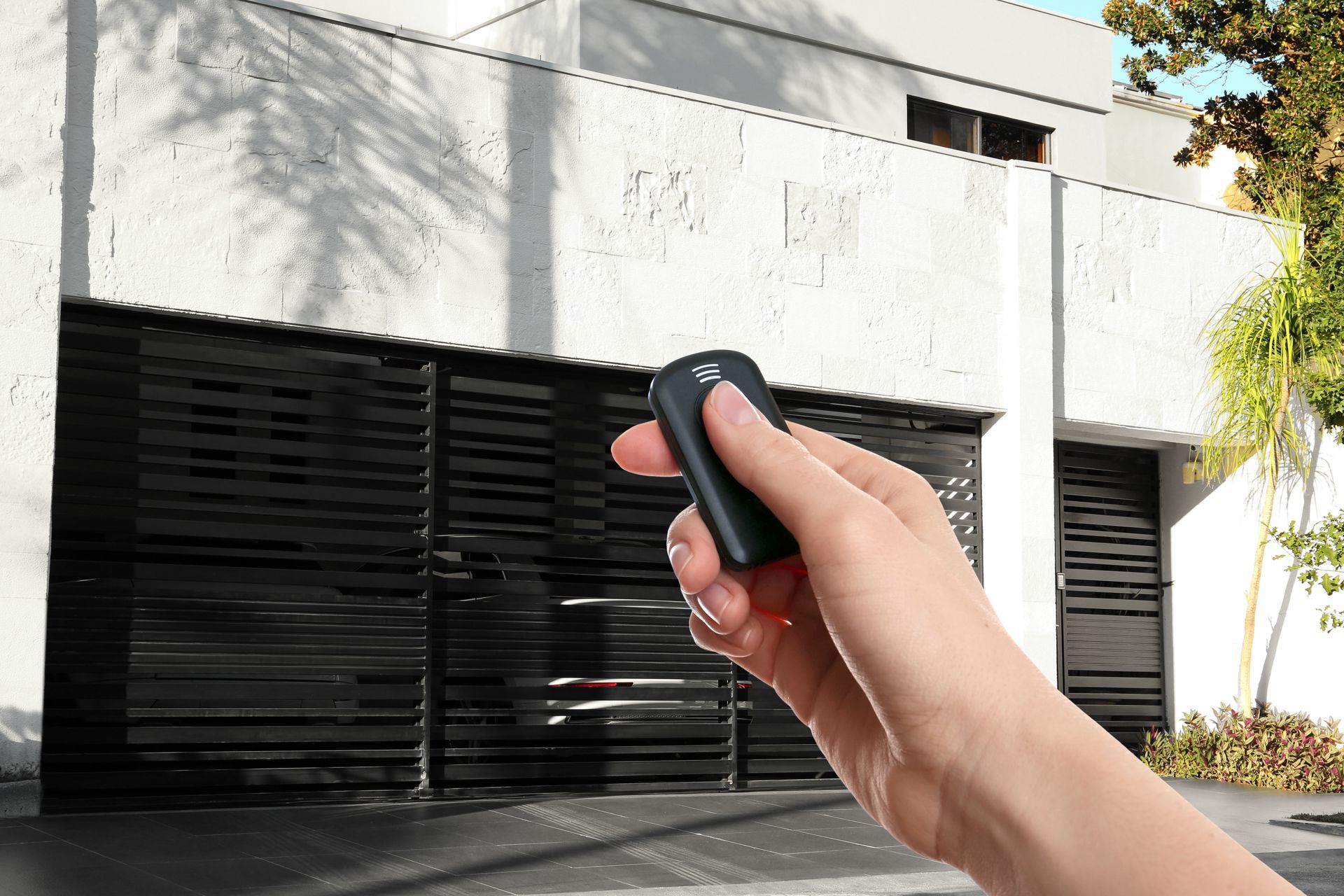 Une main tenant une télécommande noire était pointée vers une porte de garage moderne devant un bâtiment blanc.
