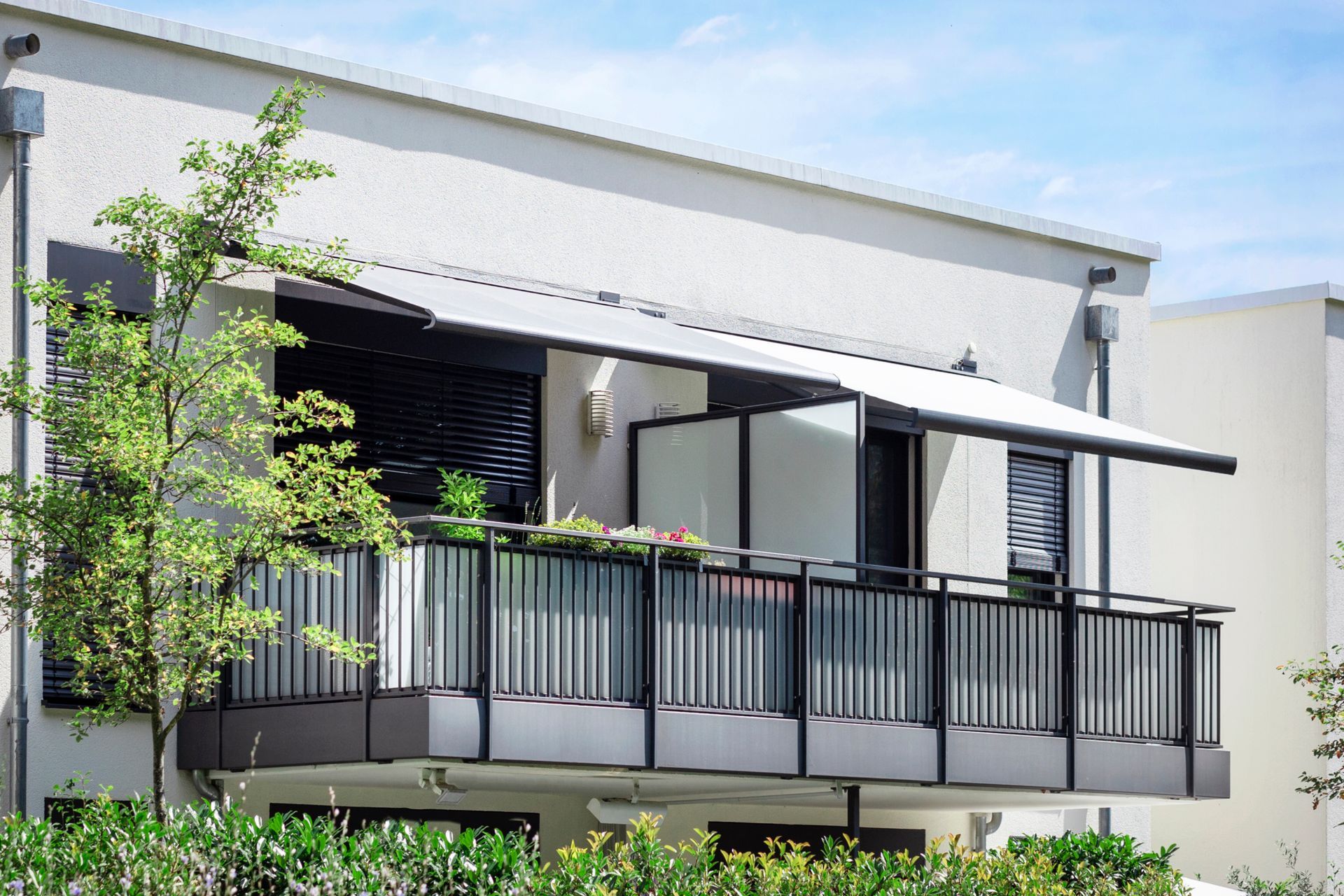 Un balcon moderne avec une rambarde en métal noir et un store rétractable en tissu blanc, sur la façade d'un bâtiment beige.