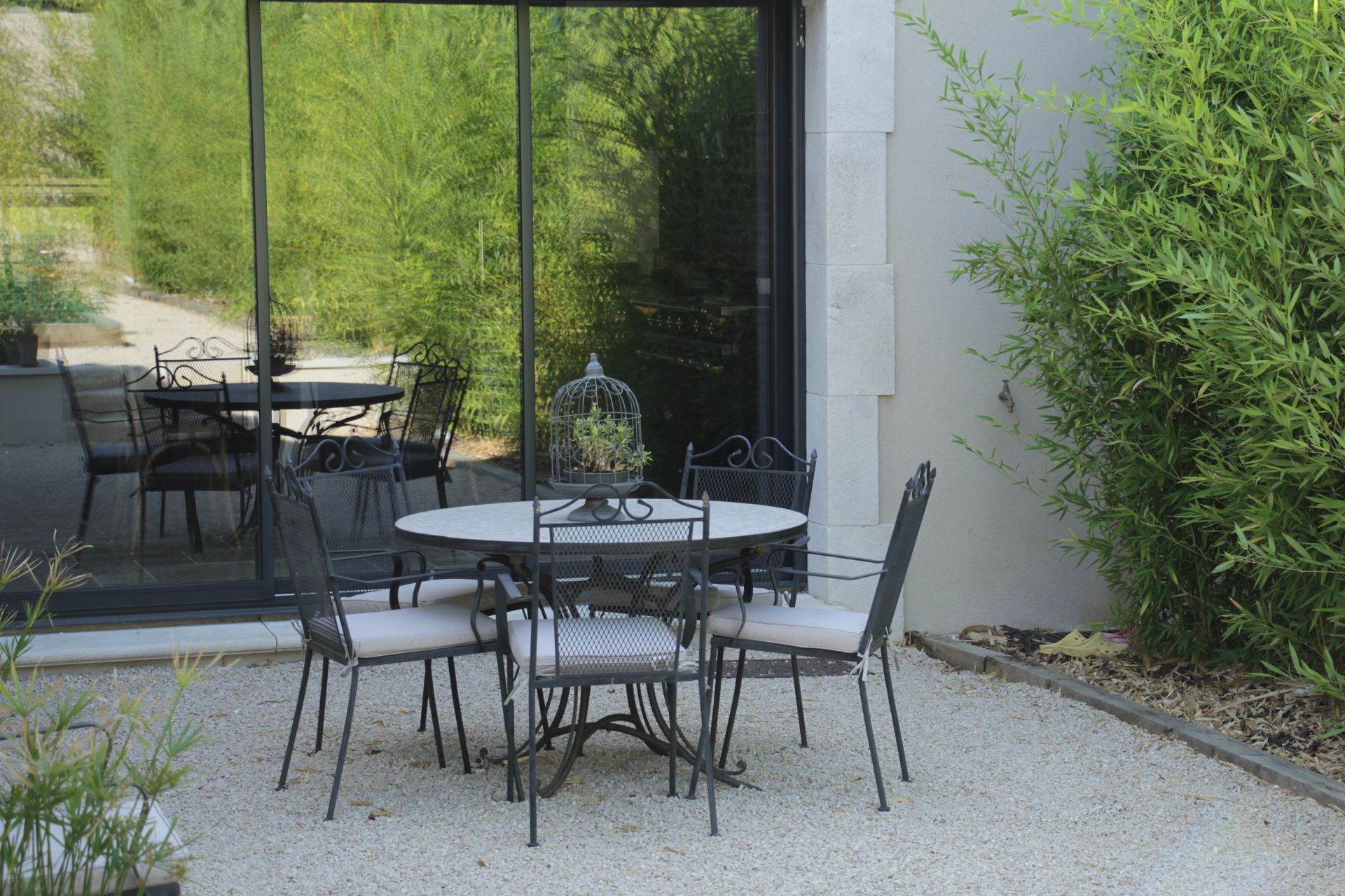 Une table ronde et quatre chaises de jardin sont installées sur une terrasse en gravier, à côté d'une porte coulissante.