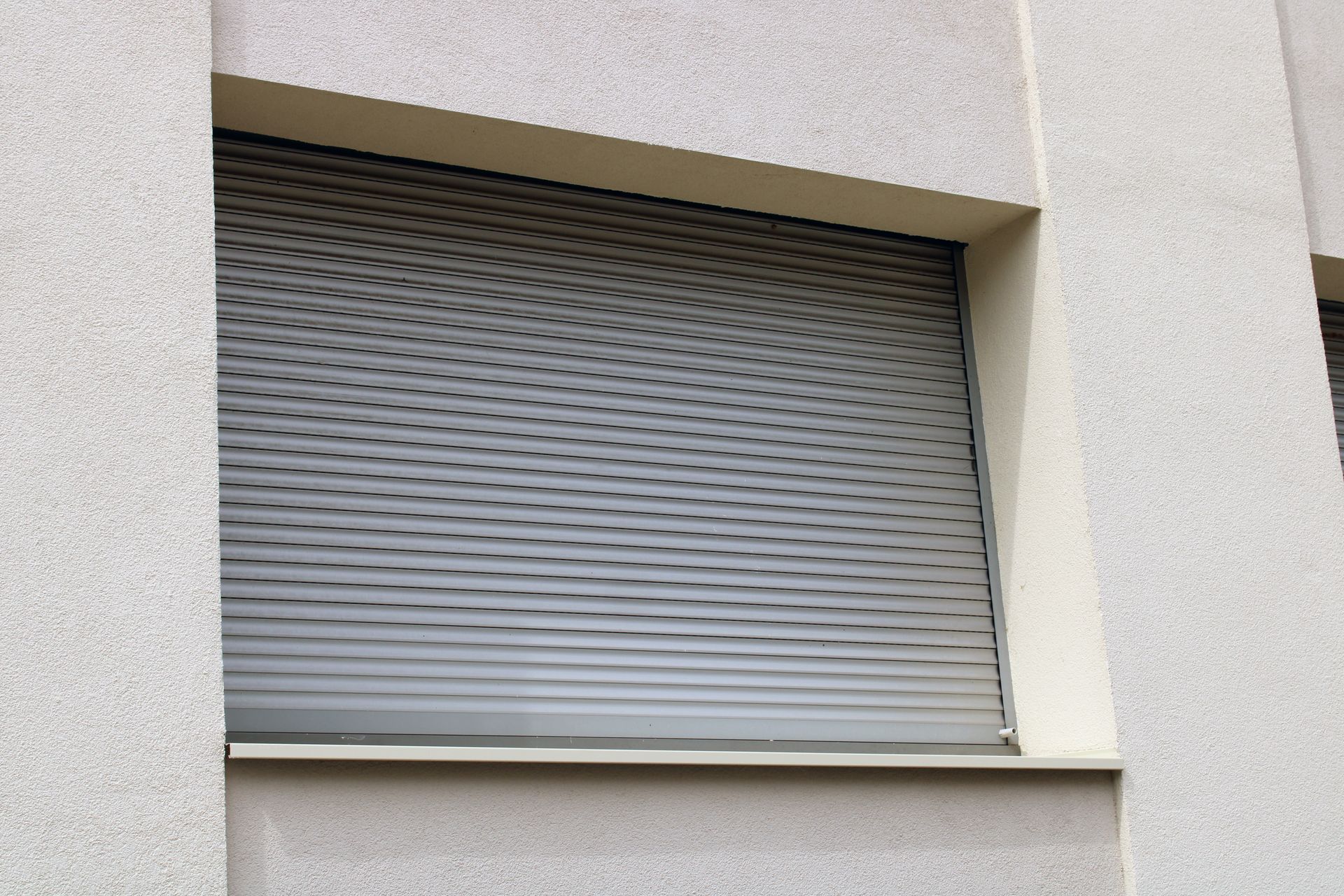 Un volet roulant gris fermé recouvre une fenêtre rectangulaire sur un mur extérieur texturé de couleur claire.
