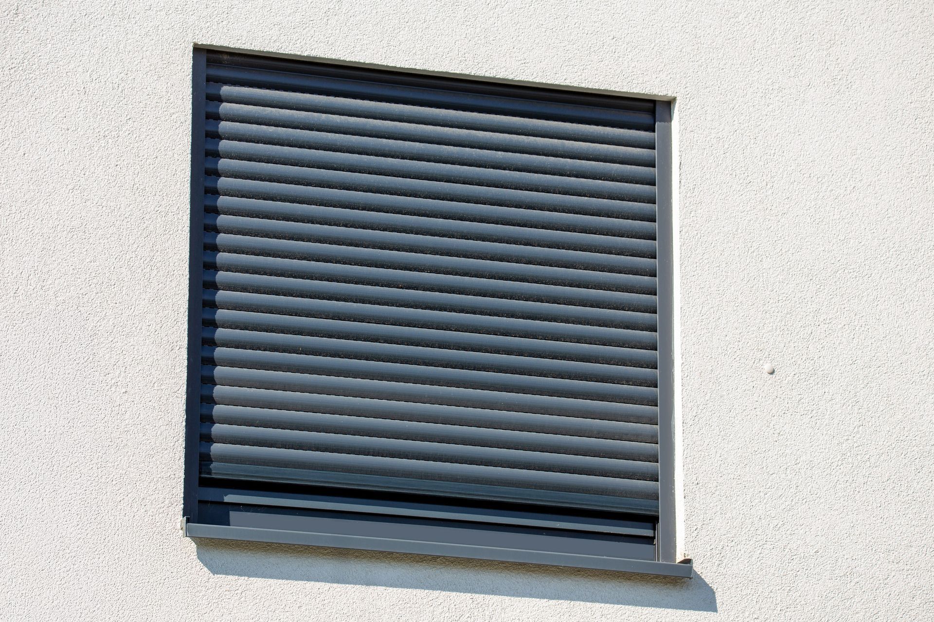 Une fenêtre rectangulaire sur un mur blanc texturé, recouverte par des volets roulants extérieurs gris foncé.
