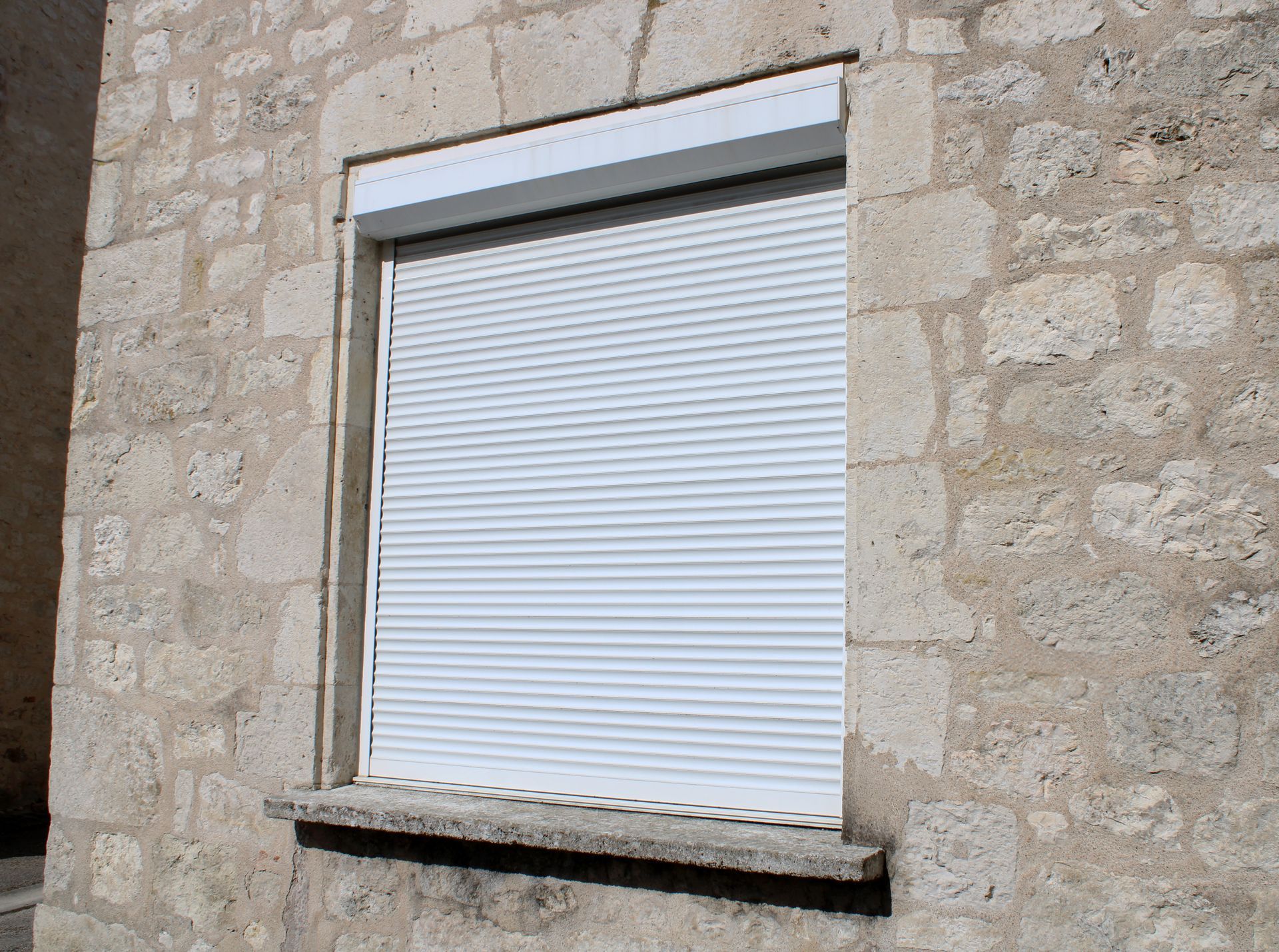 Un volet roulant blanc fixé sur un mur en pierre texturée de couleur claire.