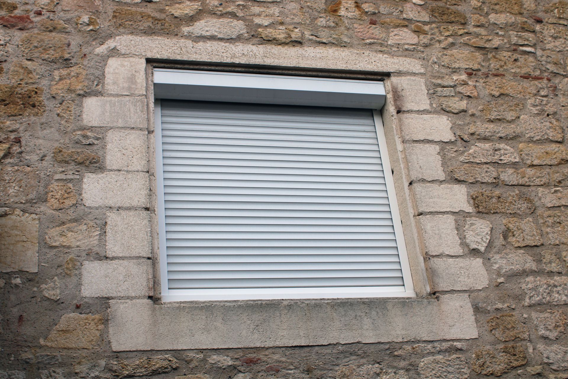 Une fenêtre aux volets gris clair fermés, encastrée dans un mur de pierre rustique.