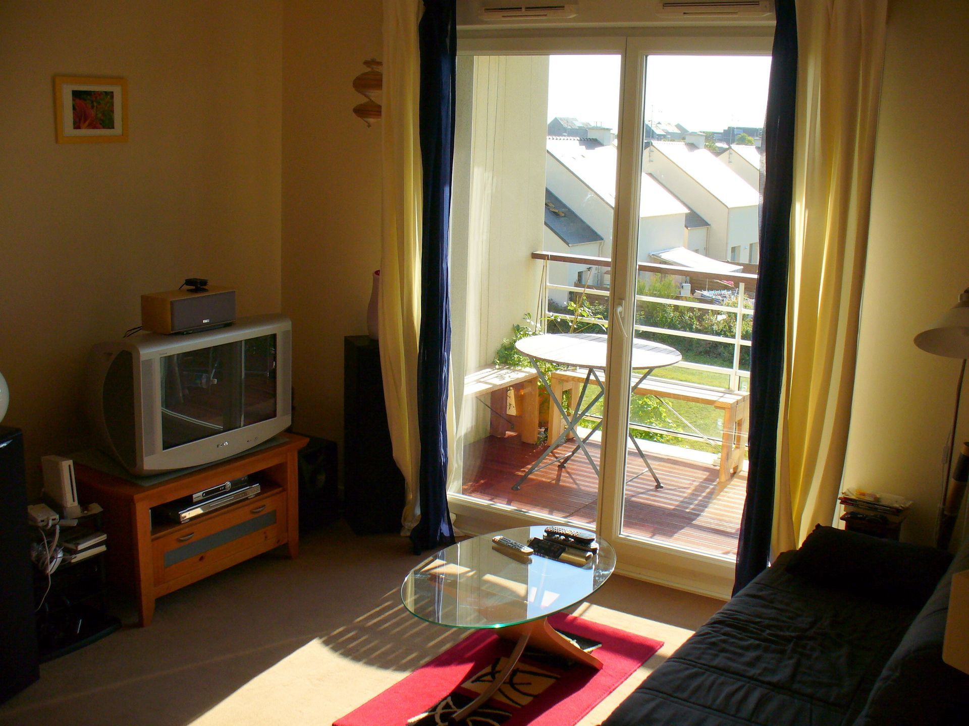 Un salon avec une télévision, une table basse, un tapis rouge et une porte vitrée donnant sur un balcon.