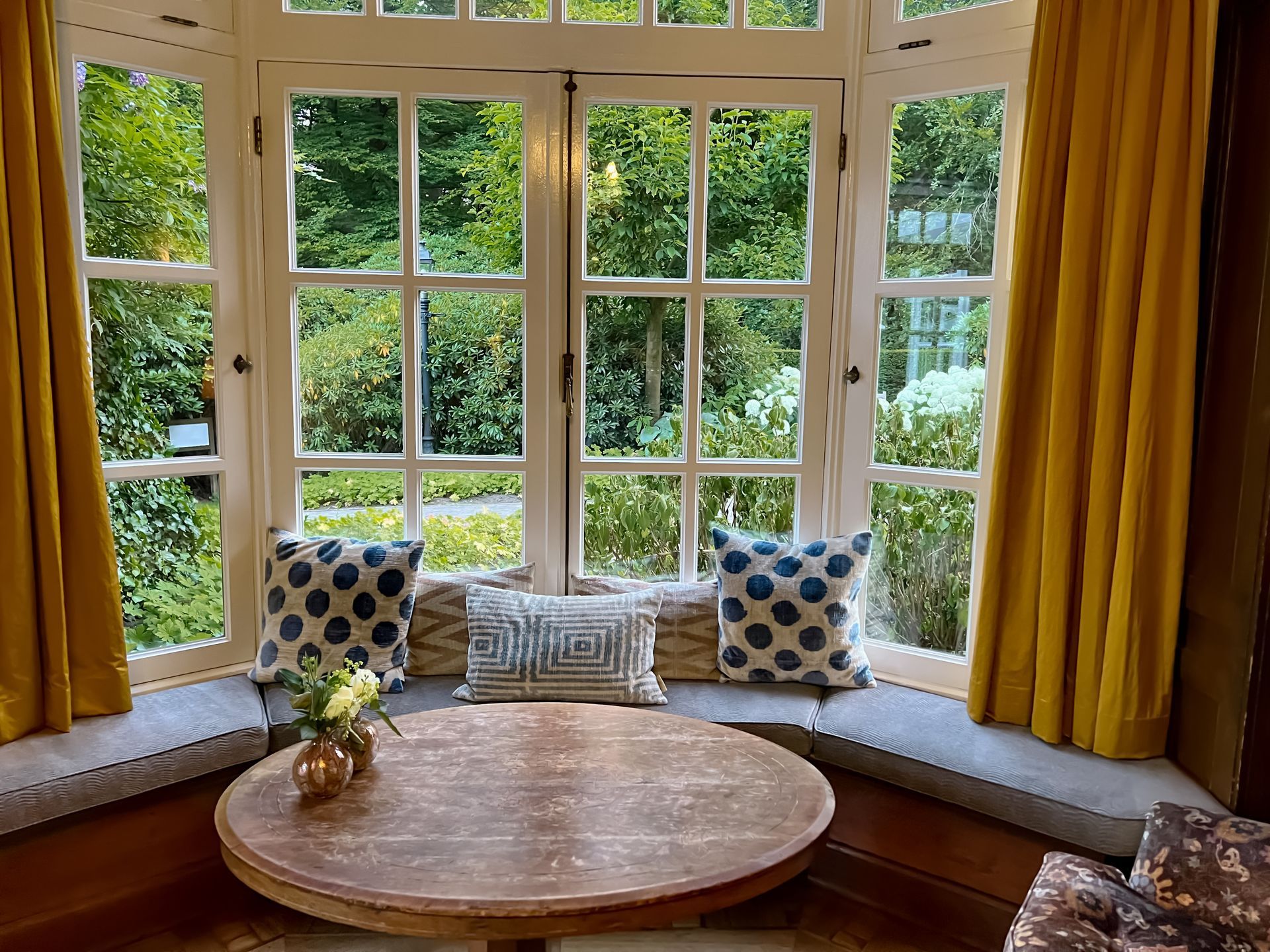 Un coin salon sous une baie vitrée avec des rideaux moutarde, une table ronde en bois et des coussins décoratifs à motifs.