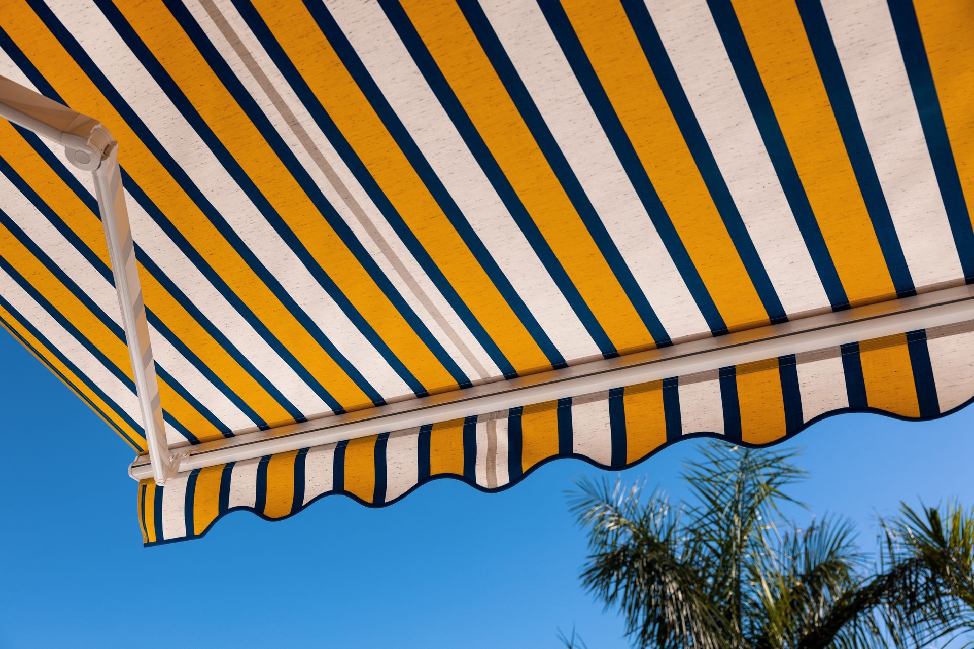 Un store rétractable à rayures jaunes et bleu foncé sous un ciel bleu clair, avec des palmes de palmier visibles en bas.