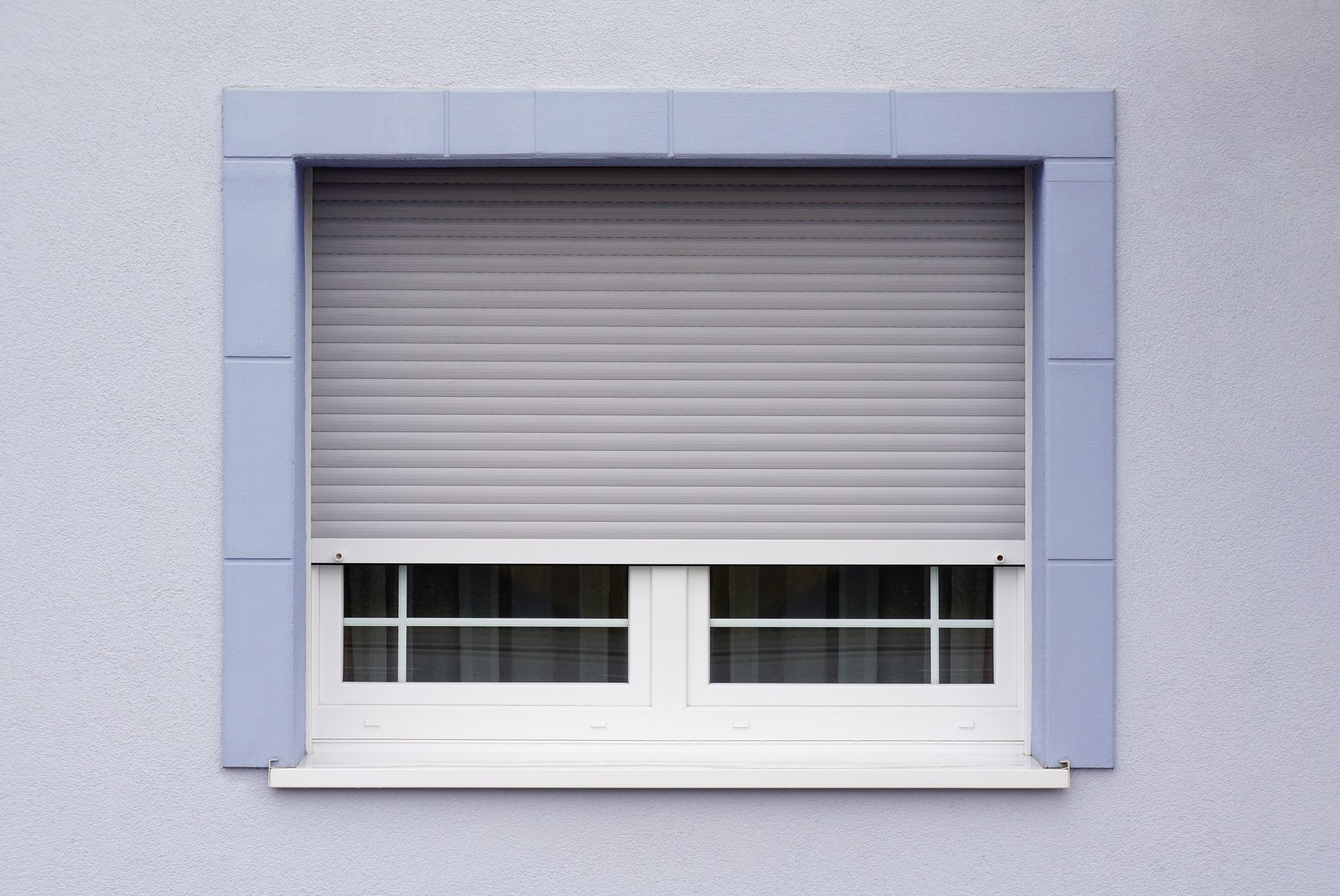 Une fenêtre à cadre blanc et volet roulant gris partiellement fermé, adossée à un mur à la texture claire.
