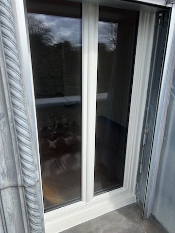 Porte-fenêtre coulissante en verre dans un mur extérieur à bardage gris, reflétant les arbres et la balustrade de la terrasse.