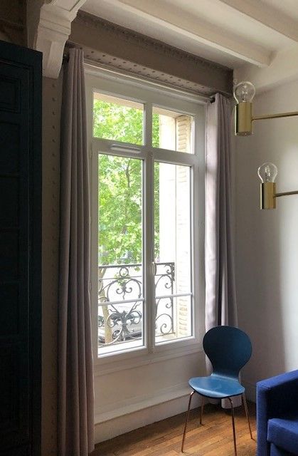 Pièce lumineuse avec une grande fenêtre, des rideaux blancs, une chaise bleue et un canapé foncé.