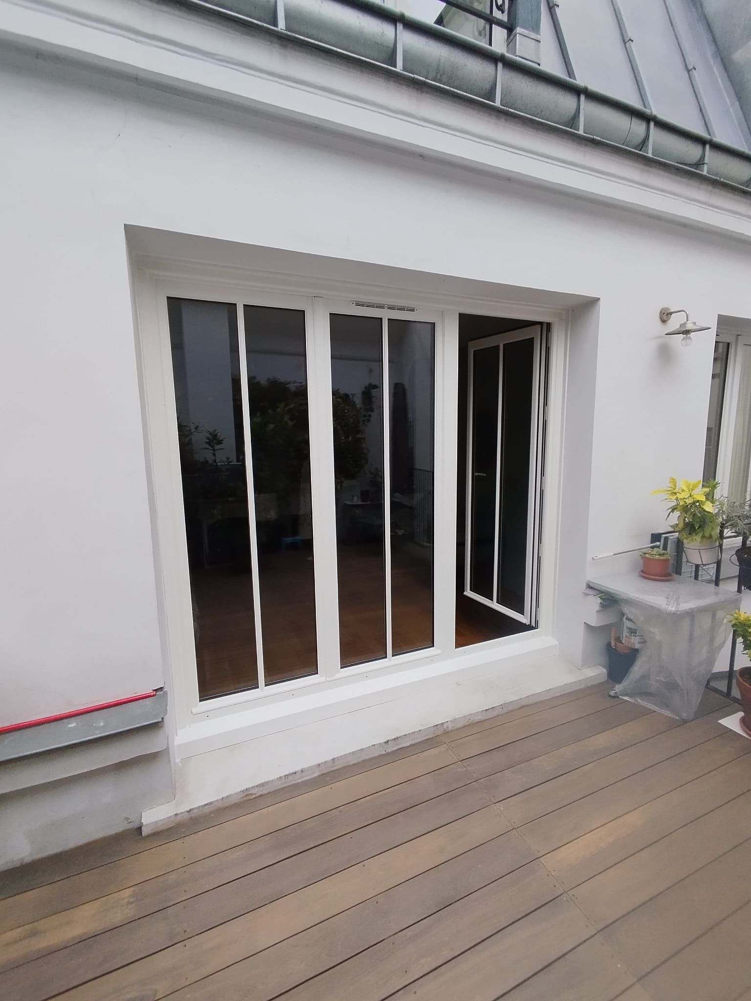 Mur extérieur blanc avec une porte coulissante en verre donnant sur une terrasse en bois sur le toit agrémentée de plantes en pot.