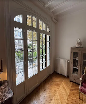 Haute fenêtre cintrée à encadrement blanc dans une pièce avec parquet à chevrons, radiateur et armoire vitrée.