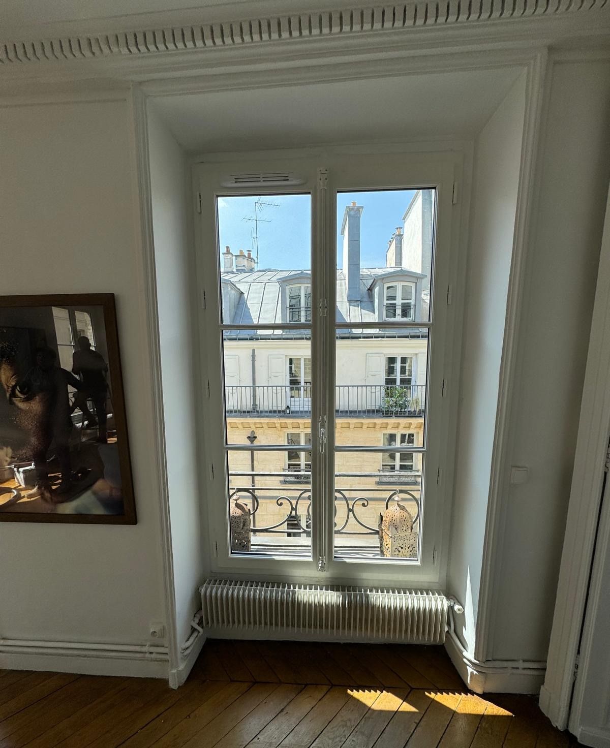 Haute fenêtre dans une pièce blanche donnant sur les immeubles parisiens et le ciel bleu.