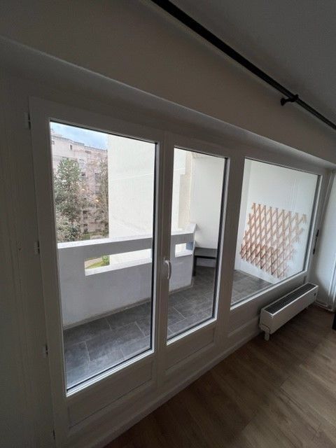 Vue sur balcon à travers des portes vitrées à cadre blanc dans une pièce lumineuse, avec parquet et radiateur.