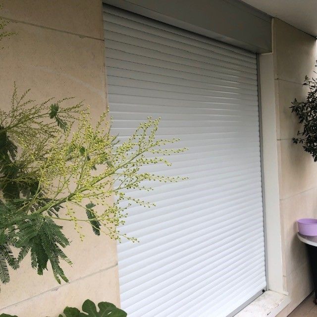 Volet roulant blanc fermé sur l'entrée d'un immeuble beige, avec de la verdure au premier plan.