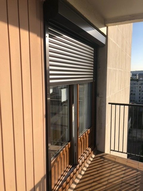Un volet roulant marron est partiellement abaissé sur une porte vitrée et une fenêtre adjacente, sur un mur extérieur blanc.