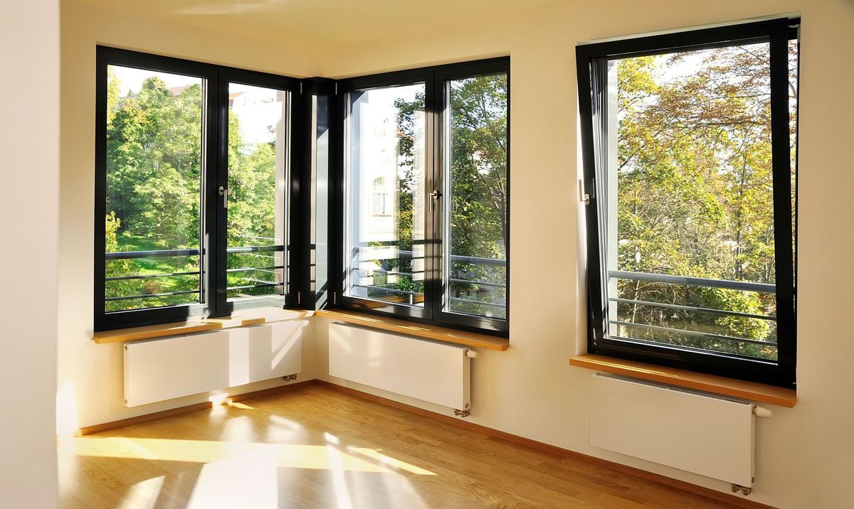 Chambre d'angle lumineuse avec parquet, radiateurs blancs sous fenêtres à cadre noir et vue sur les arbres à l'extérieur.