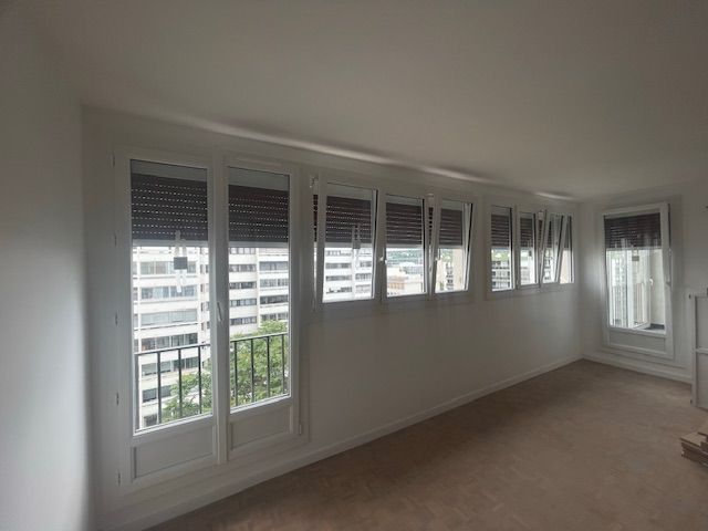 Pièce lumineuse et vide avec de hautes fenêtres, des murs blancs, une moquette beige et une porte miroir.