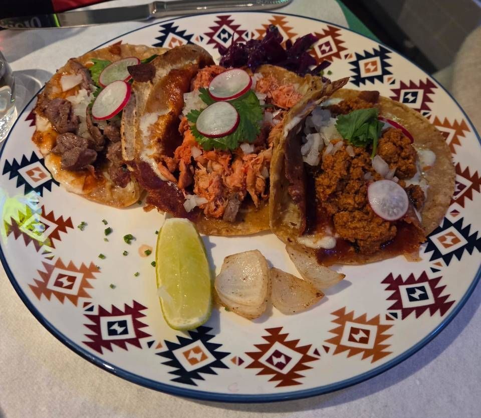 Drei Tacos auf einem gemusterten Teller mit Limettenvierteln und Radieschenscheiben, garniert mit Koriander und Salsa.