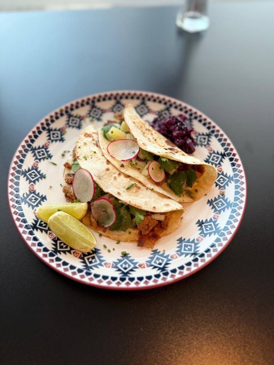 Gemusterter Teller mit einem Taco, belegt mit Radieschenscheiben und Kräutern, serviert mit einer Limettenspalte auf einem dunklen Tisch